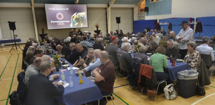 Stemningsbillede fra Foreningernes Aften. Folk sidder ved langborde og for enden er en storskærm, hvorpå der står velkommen. Foto: Kim Haugaard